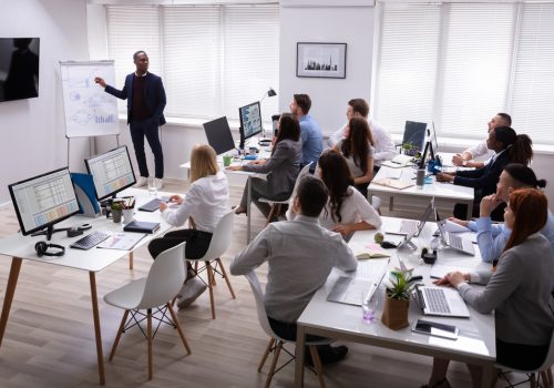 African Businessman Giving Presentation To His Colleagues Sitting On Table With Computers In Office